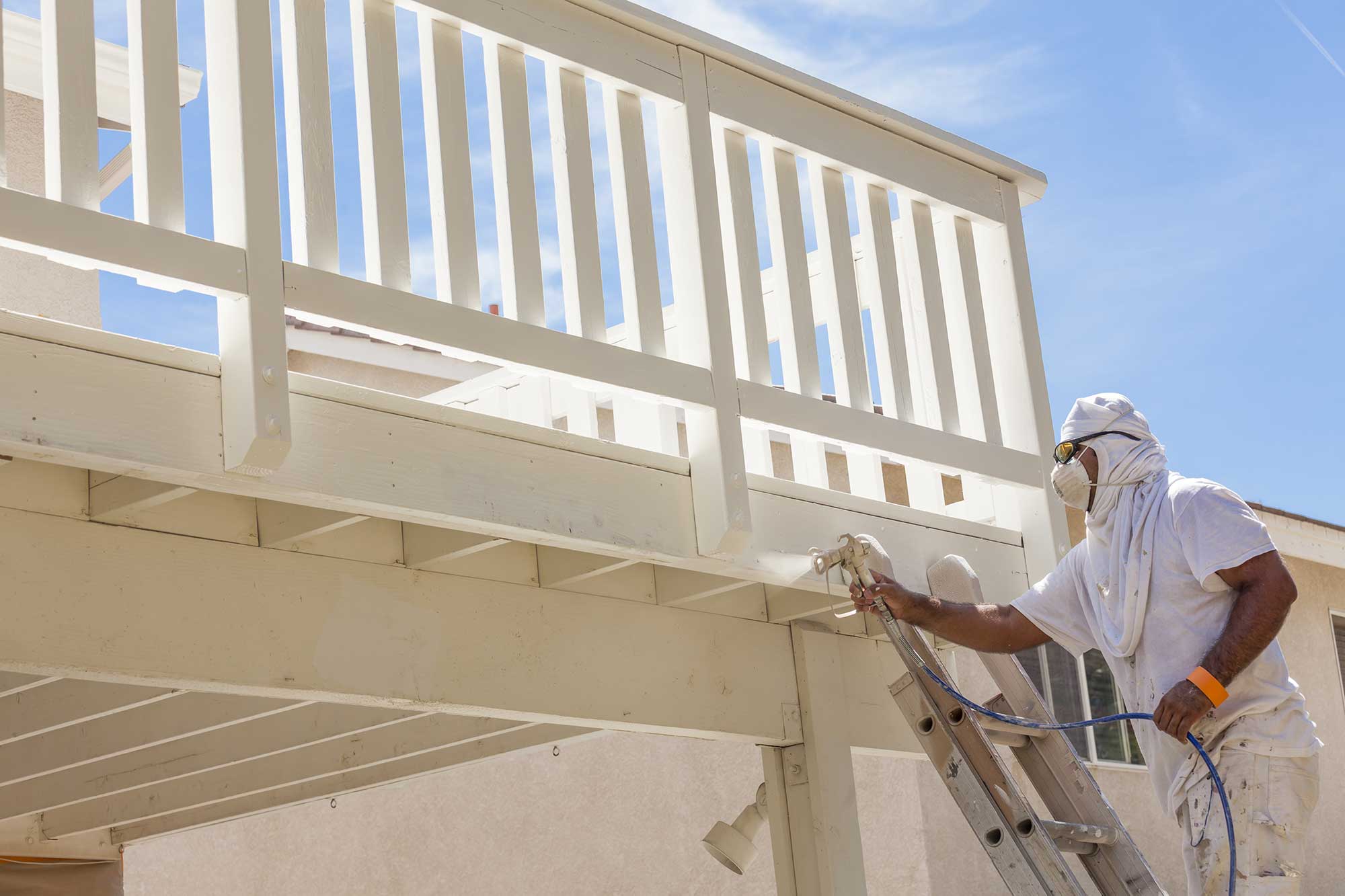 A person wearing safety gear, including goggles and a mask, stands on a ladder while spray-painting a white wooden railing under a clear blue sky.