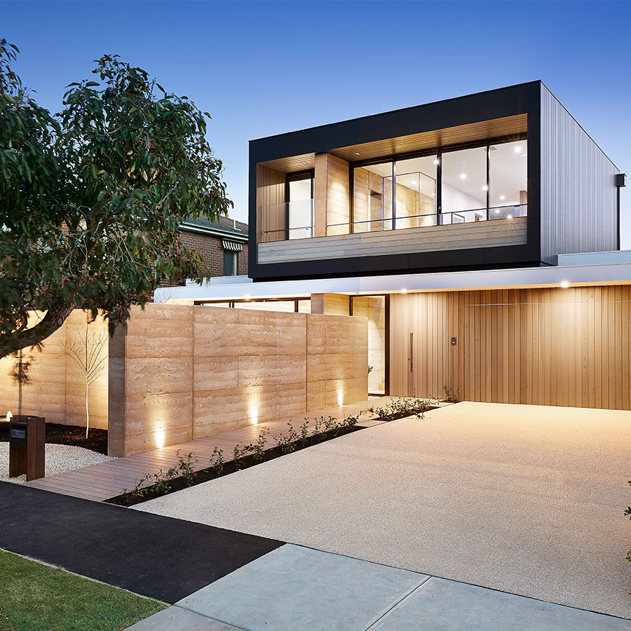 Modern two-story home with a mix of wood and stone, illuminated pathway, and a landscaped yard under a clear blue sky.