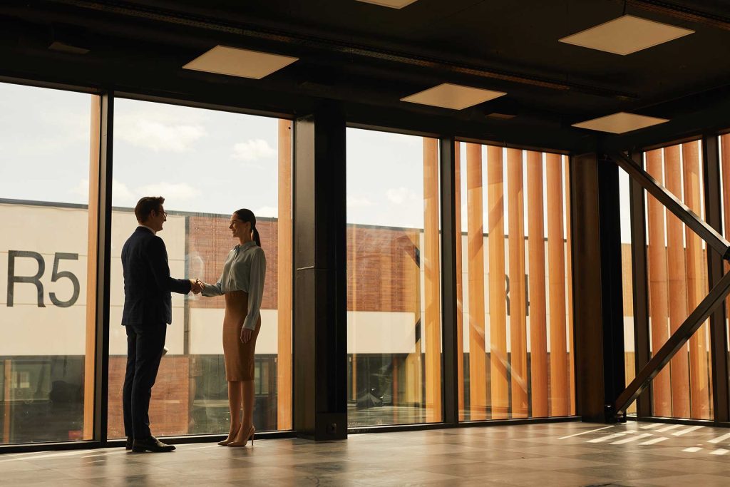 A man in a suit shakes hands with a woman in a blouse and skirt inside a modern office space with large windows.