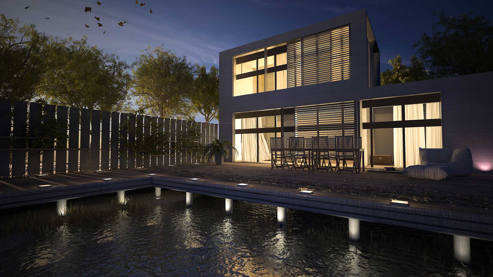 A modern two-story house with large windows, outdoor dining area, and a wooden deck over a reflective pool, surrounded by trees at dusk.