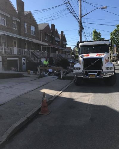 Construction scene on a residential street, featuring a large truck and workers near a pile of debris, with houses and power lines in the background.