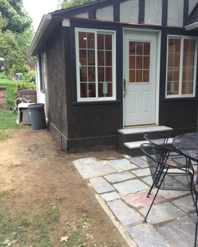 Exterior view of a small house with a white door, large windows, and a stone patio with black chairs. A trash can is nearby.