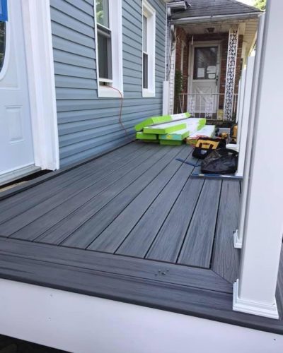 A new gray composite deck is under construction beside a blue-sided house, with materials and tools scattered nearby.