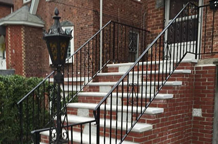 Black iron railing surrounds a set of white stone steps leading to a brick house, with a decorative lamp by the entrance.