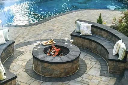 A cozy outdoor space with a circular stone fire pit, surrounded by curved seating and a pool in the background, pillows on chairs.